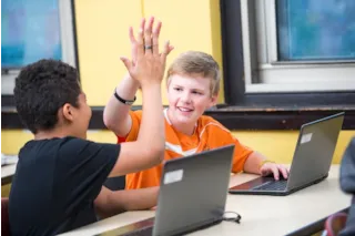 Two students in a classroom high-fiving while sitting at a table with laptops.