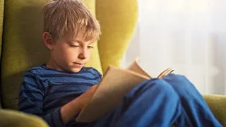Young boy sitting in a green chair, wearing blue stripes, reading a book with sunlight in the background.
