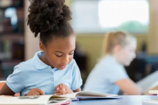 Young girl in a blue shirt reading a book at a desk in a classroom, with another student in the background.