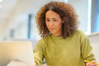 Woman with curly hair in a green sweater working on a laptop and holding a pencil in a bright office.