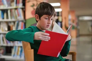 A boy in a green shirt sits in a library, reading a red book with shelves of books in the background.