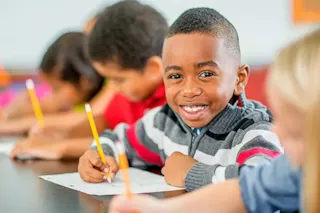 Smiling boy in a striped sweater writes with a pencil in class, surrounded by other children at a desk.