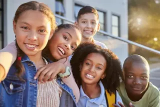 Five smiling kids pose together outdoors in sunlight, looking at the camera with a building in the background.