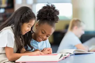 Two young girls smile and read a book together at a table, with another student reading in the background.