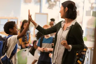 A teacher gives a high-five to a smiling student in a classroom with other kids in the background.
