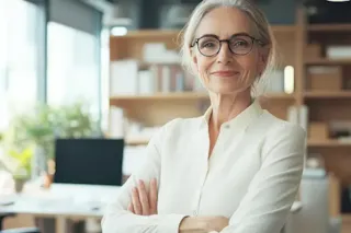 Confident older woman with glasses and gray hair stands smiling in a modern office, arms crossed.