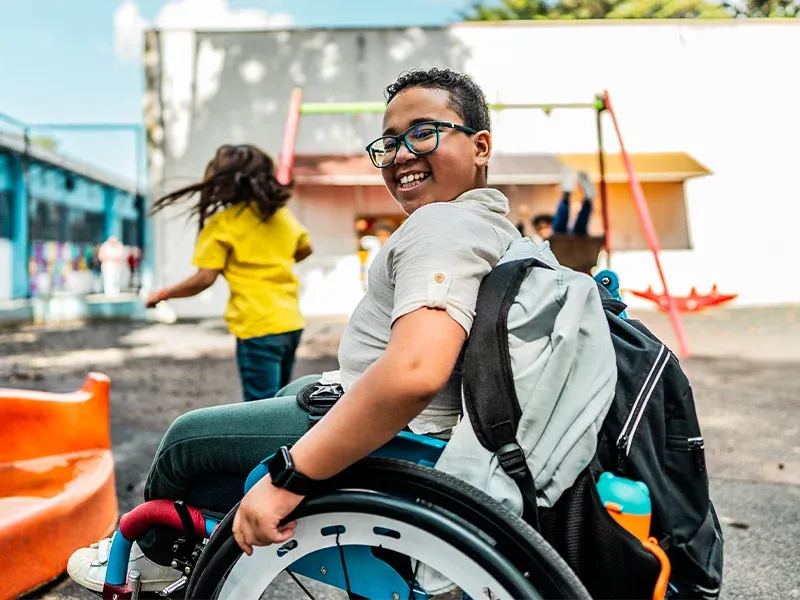 Smiling boy in a wheelchair on a playground, wearing glasses and a backpack, with children playing in the background.