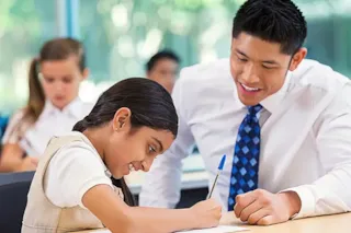 Smiling teacher helps a young girl with homework at a classroom desk, while other students work in the background.