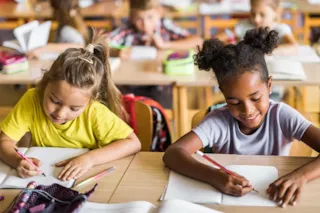 Two young girls sitting at a desk in a classroom, smiling and writing in notebooks, with other kids in the background.