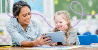 Woman and young girl smiling together while looking at a tablet in a classroom setting.