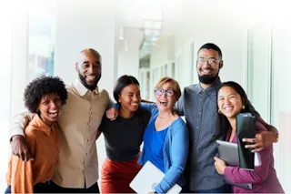 Six diverse colleagues stand in a hallway, smiling and posing closely together, holding laptops and folders.