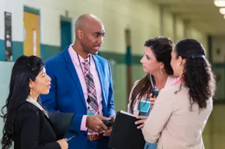 Four professionals in business attire have a serious discussion in a hallway, holding folders and a walkie-talkie.