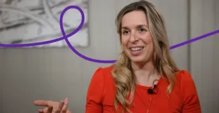 Smiling woman in a red shirt gestures while talking, with a purple line looping behind her on the wall.