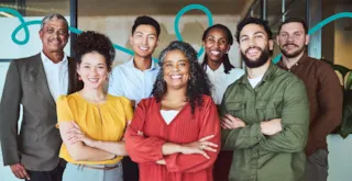 Seven diverse people standing together smiling in an office setting, arms crossed, facing the camera.