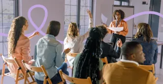 A woman leads a discussion with a group seated on chairs; one participant raises her hand to speak.