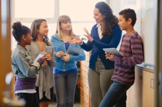 Teacher and four students standing in a classroom, holding tablets, engaged in a discussion near a whiteboard.