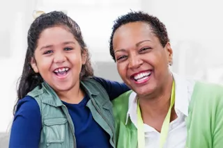 Smiling woman and young girl with dark hair and a bow, both laughing. The woman is wearing a green sweater.