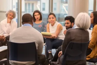 A diverse group of people sitting in a circle for a discussion in a bright room with large windows.