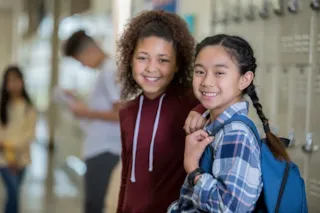 Two smiling girls stand by lockers in a school hallway, one with a backpack and the other wearing a hoodie.