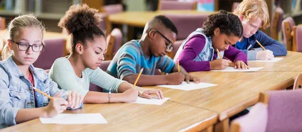Five children sit at a table in a library, focused on writing on sheets of paper with pencils.