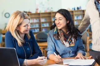 Two women sitting at a table in a library, smiling and discussing papers. One holds a pen, the other gestures with her hand.