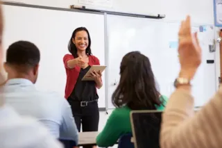 Smiling teacher holding a tablet points at a student raising their hand in a classroom with a whiteboard.