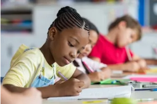 Young girl with braided hair focuses on writing in a notebook, with other children working in the background.
