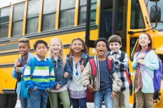 Seven smiling children with backpacks stand in front of a yellow school bus on a sunny day.