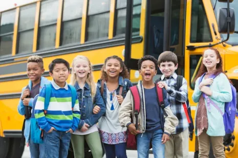 Seven smiling children with backpacks stand in front of a yellow school bus, ready for school.