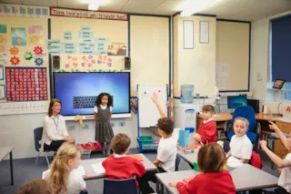 A girl stands near a smartboard in a classroom as children in red uniforms sit and raise their hands.