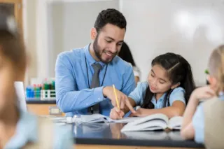 A teacher helps a young student with her work at a classroom desk, both smiling and engaged.