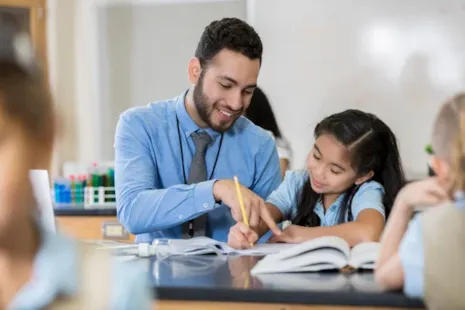 A teacher smiles while helping a young student with her work at a classroom desk.