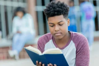 Teenager with short curly hair sits outside and reads a book, with blurred people in the background.