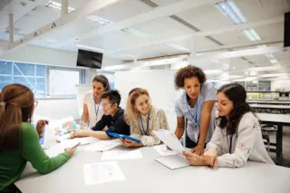 A group of students and a teacher collaborate at a table in a bright, modern classroom.