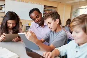 A teacher helps three students using tablets at a classroom table, all focused and engaged with their devices.
