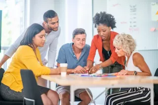 Five people gather around a table, collaborating and discussing documents in a modern office setting.
