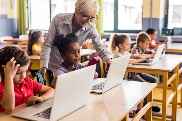 A teacher helps a young student with a laptop in a classroom of children using computers at their desks.