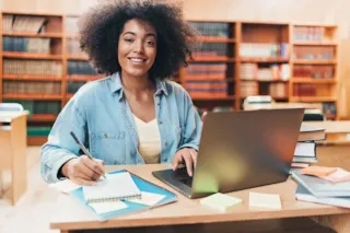 A person smiling at a library desk, using a laptop and taking notes, surrounded by books and sticky notes.