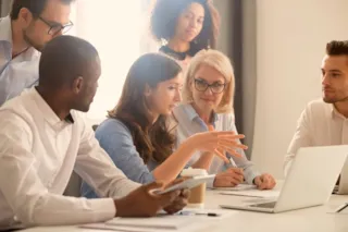 Diverse group of colleagues in a meeting room, discussing around a table with a laptop, notes, and coffee cup.