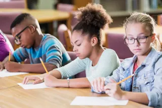 Three students sit at a table, focused on writing with pencils on paper in a classroom or library setting.