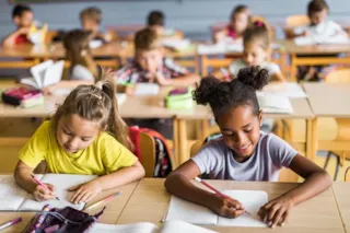 Young children sitting at desks in a classroom, smiling and writing in notebooks.