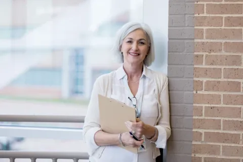 Smiling woman with gray hair, holding a clipboard, stands against a brick wall near a window.