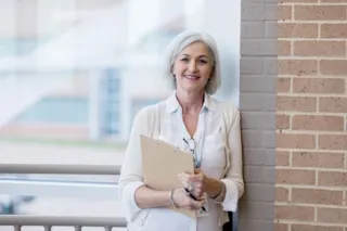 Smiling older woman with gray hair holds a clipboard, standing indoors by a brick wall and window.