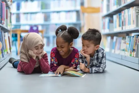 Three young children lie on the library floor, smiling and reading a book together between bookshelves.