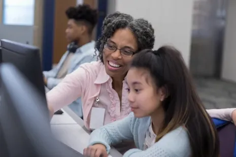 Smiling teacher helps a young student on a computer in a classroom with other students in the background.