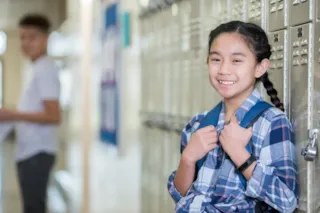 A student with braided hair smiles while standing by lockers in a hallway. Another student is in the background.