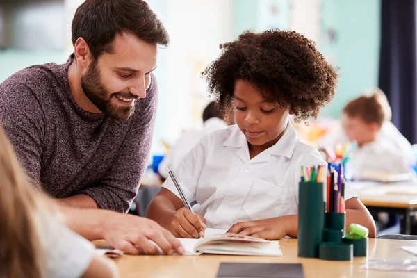 A teacher helps a young student with schoolwork at a classroom desk, both smiling and focused on the task.