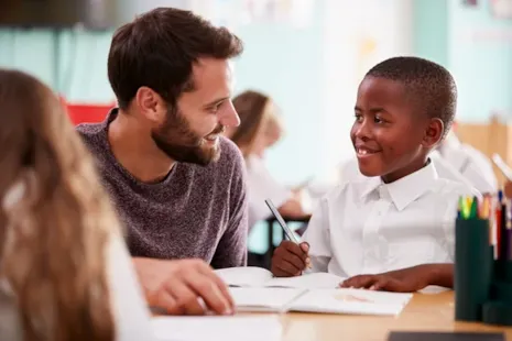 Teacher and student smiling and talking at a desk in a classroom, with open books and pencils in front of them.