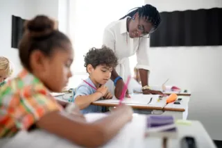 A teacher helps a young student with classwork while other children work at their desks in a classroom.