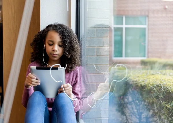 A young girl with curly hair sits by a window, using a tablet and wearing earphones, appearing focused.